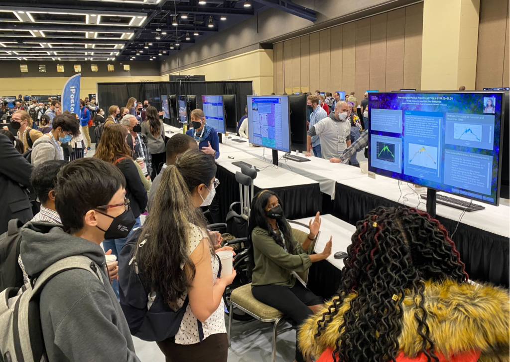 An image from the iPoster sessions at AAS241 Winter meeting. a large screen on a sitting desk which is displaying an iPoster. The poster presenter sits in a chair with their hands raised up towards the screen in an explanatory gesture. 