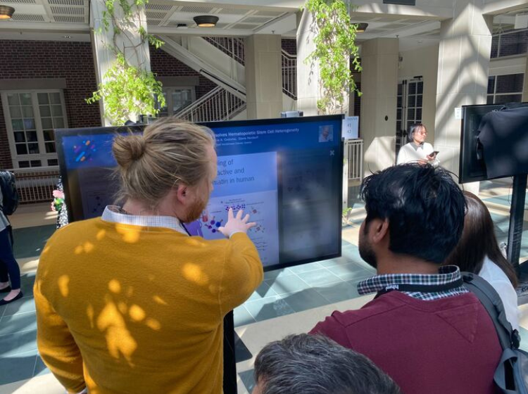 A person presenting an iPoster on a large screen to 3 people standing nearby.