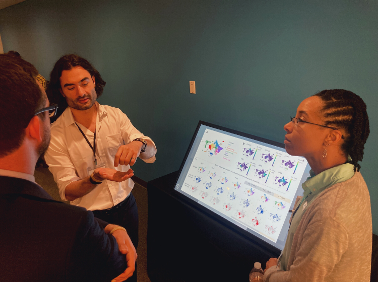 3 people standing next to an iPoster on a big screen at the V Scholar Summit 2022. One person is explaining something to the other two.