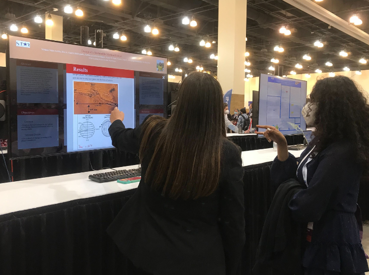 2 people at AAS240 standing in front of a large screen. One of them is touching the screen which shows an iPoster and an image of a graph. The other person is pointing towards the screen from a distance. 