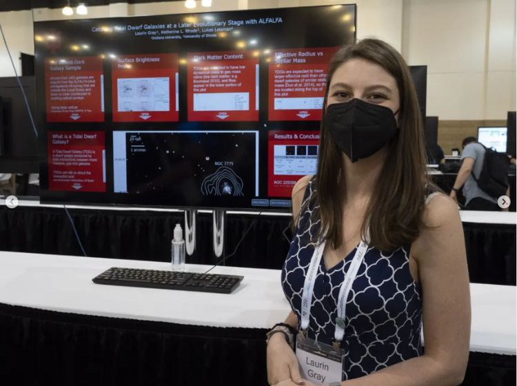 Presenter standing next to a large screen, facing the camera. On the screen is an iPoster with 6 red content boxes and a black background. 