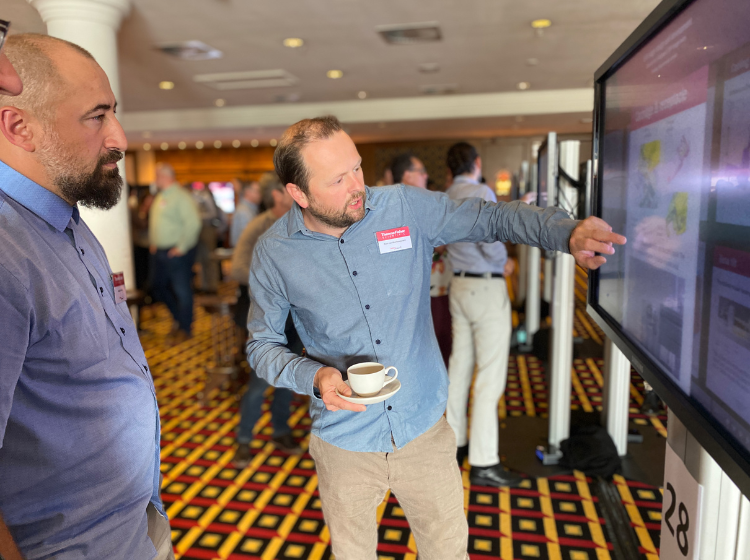 Close up of two people, both are facing a large screen with an iPoster on it. One of them is touching the screen and holding a cup and saucer. 