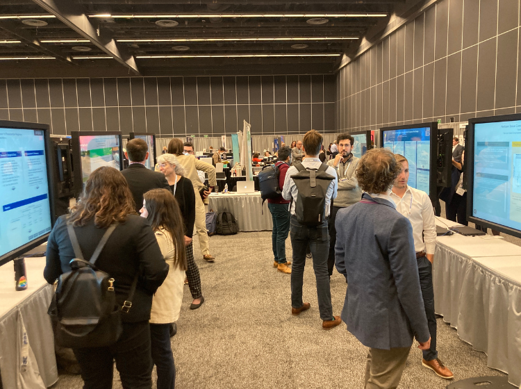Participants at the APSA annual meeting standing in pairs around 6 iPoster screens. The screens have been set up on two long tables that are facing each other. 