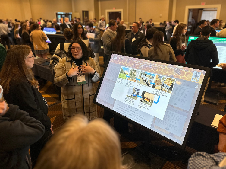 Person at K-INBRE Symposium standing to the side of a large angled screen with an iPoster on it. A group of onlookers stand around  looking at the screen. 
