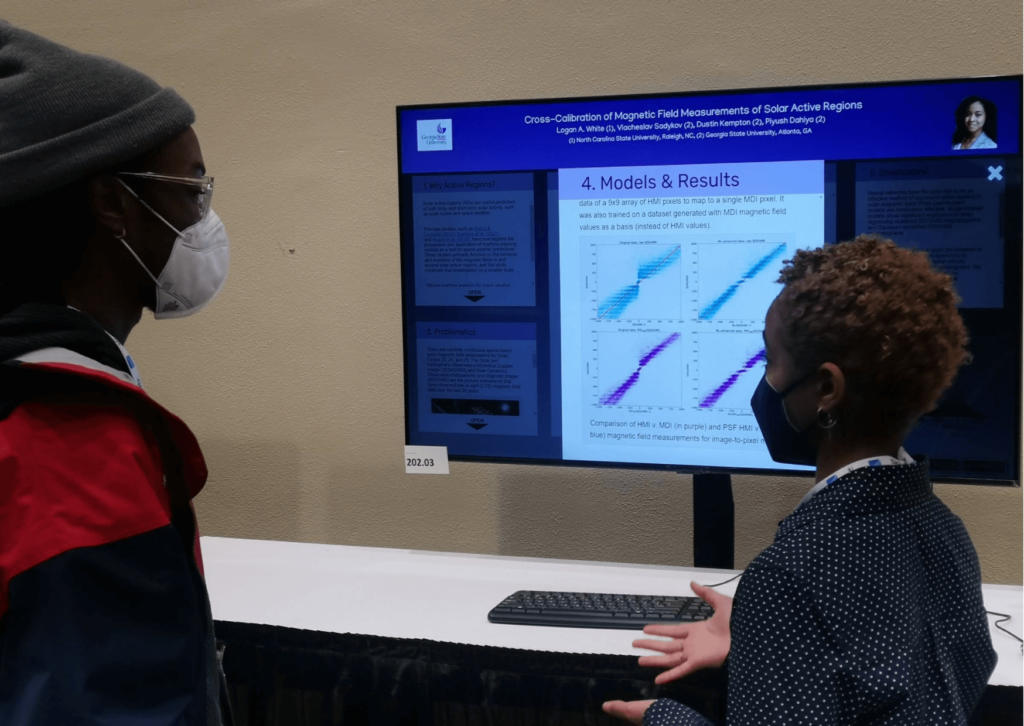 An image from the iPoster sessions at AAS241 Winter meeting. a large screen on a standing desk which is displaying an iPoster. 2 people stand wither side of the screen and they're looking at each other as one of them is holding out their hand in explanation.  