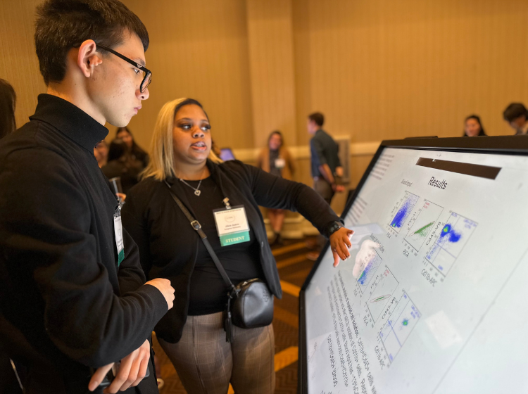 Person at K-INBRE Symposium standing in front of and touching a large angled screen with an iPoster on it. An onlooker stands beside them looking at the screen. 