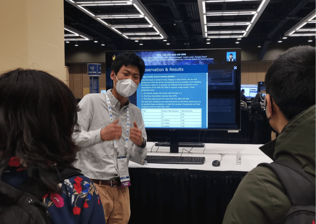 An image from the iPoster sessions at AAS241 Winter meeting. a large screen on a standing desk which is displaying an iPoster. 1 person stands infront of the screen to the left and has both thumbs up. 2 people are faced away from the camera watching the presenter. 