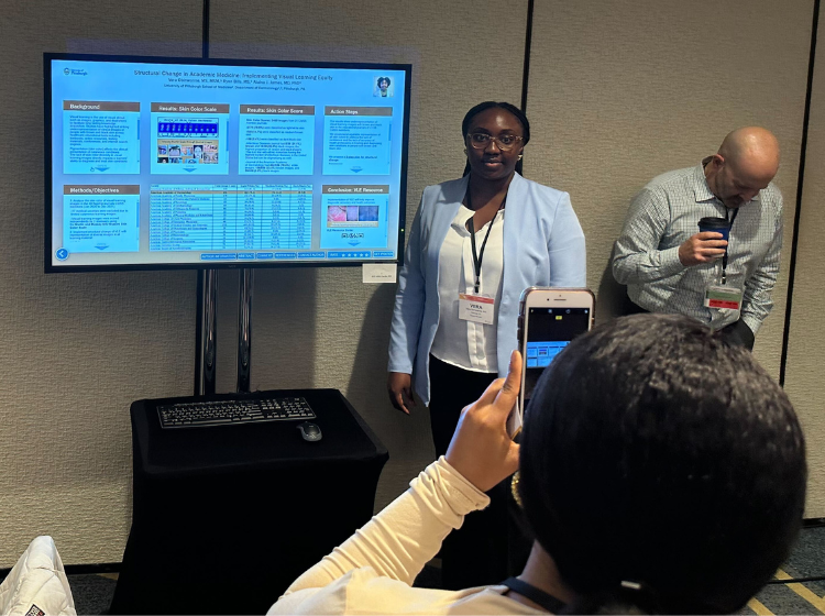 A person poses next to a screen with an iPoster displayed on it whilst another person takes a photo on a phone. 