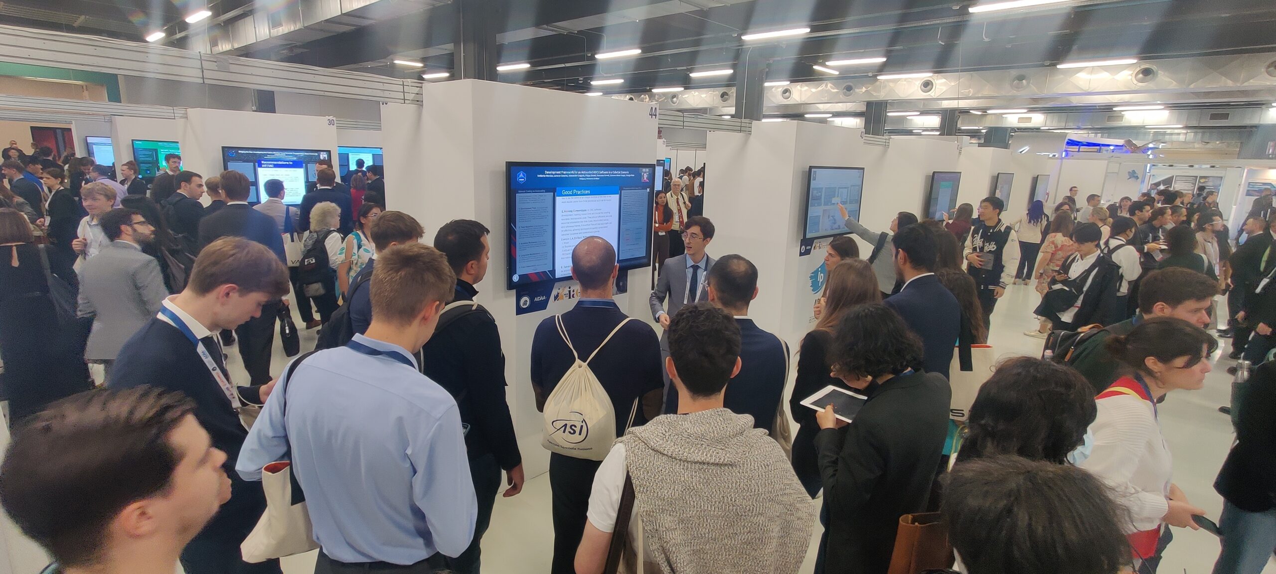 The iPoster Hall at the 2024 IAC "Responsible Space for Sustainability" Congress, in Milan, Italy. The Hall is filled with attendees listening to presentations conducted by authors on large white touch screen kiosks.