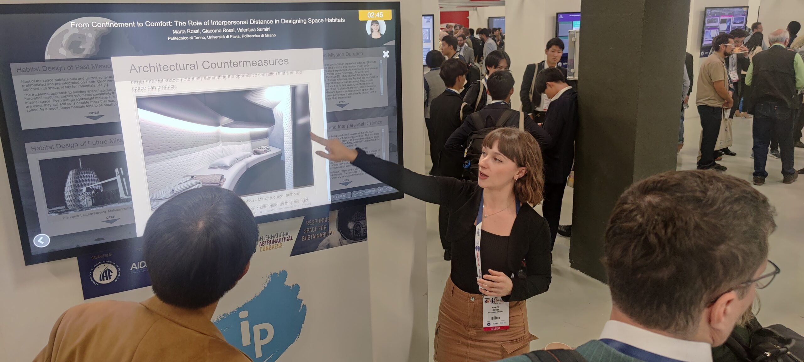 The iPoster Hall at the 2024 IAC "Responsible Space for Sustainability" Congress, in Milan, Italy. A woman is presenting her research on a large white touch screen kiosk. She is pointing at an image.