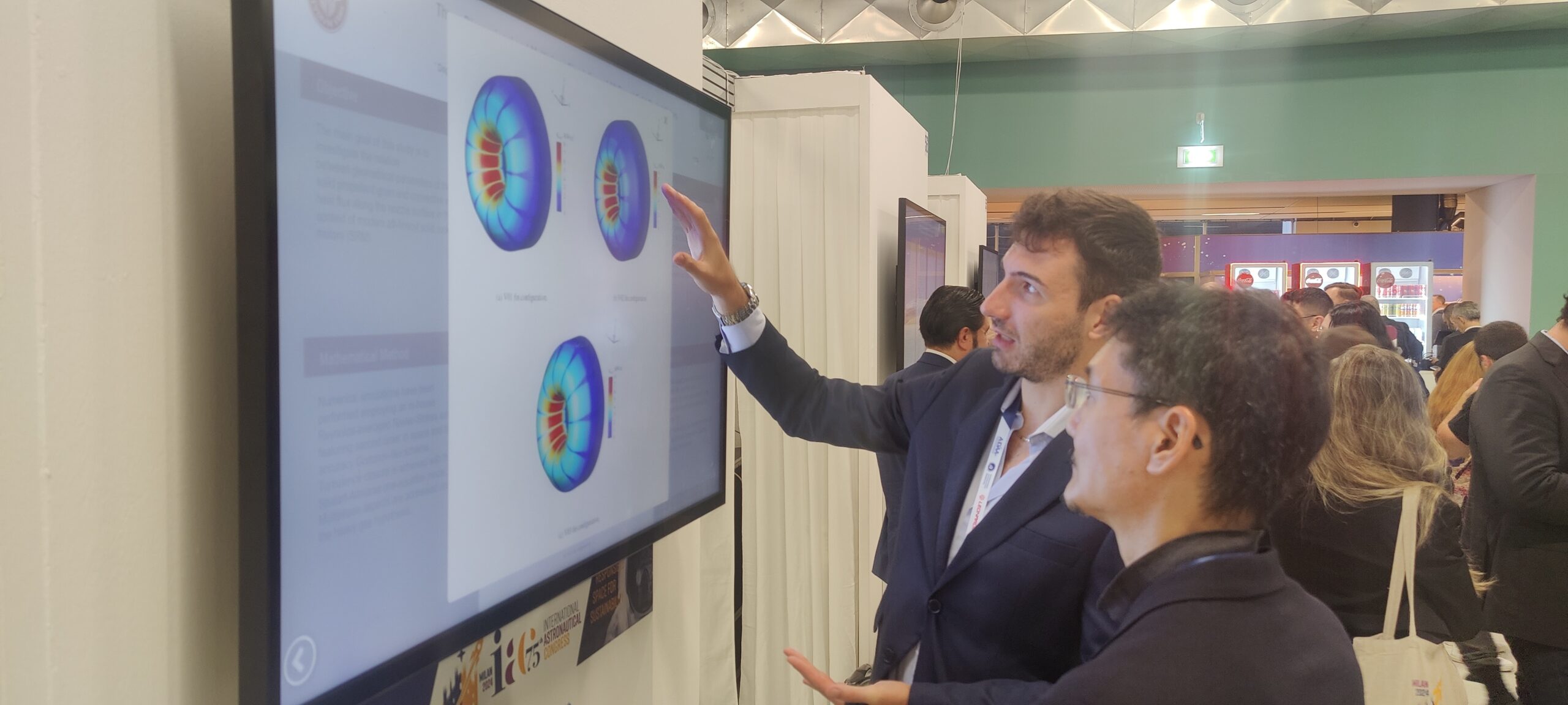 The iPoster Hall at the 2024 IAC "Responsible Space for Sustainability" Congress, in Milan, Italy. A a man is presenting his research on a large white touch screen kiosk. He is pointing at an image, explaining things to another an standing beside him.