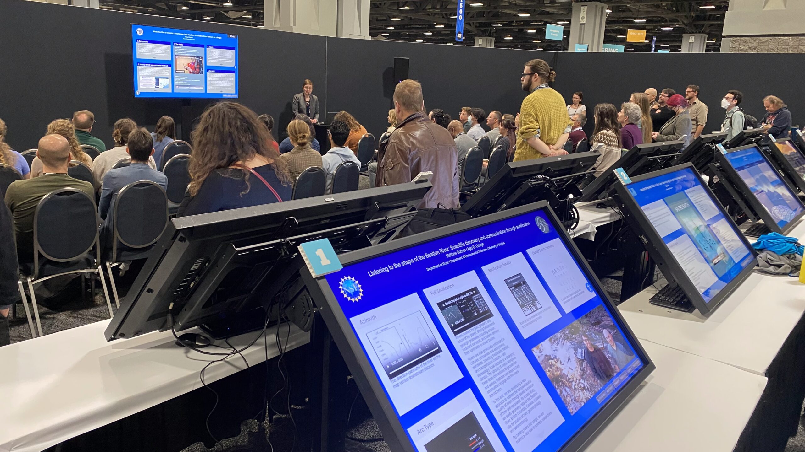 eLightning theater at AGU 2024 annual meeting. In the background a presenter giving an oral presentation on a big screen in front of a large crowd, sitting and standing. In the foreground, a row of large format touch screens. The AGU meeting theme is What's next for Science?