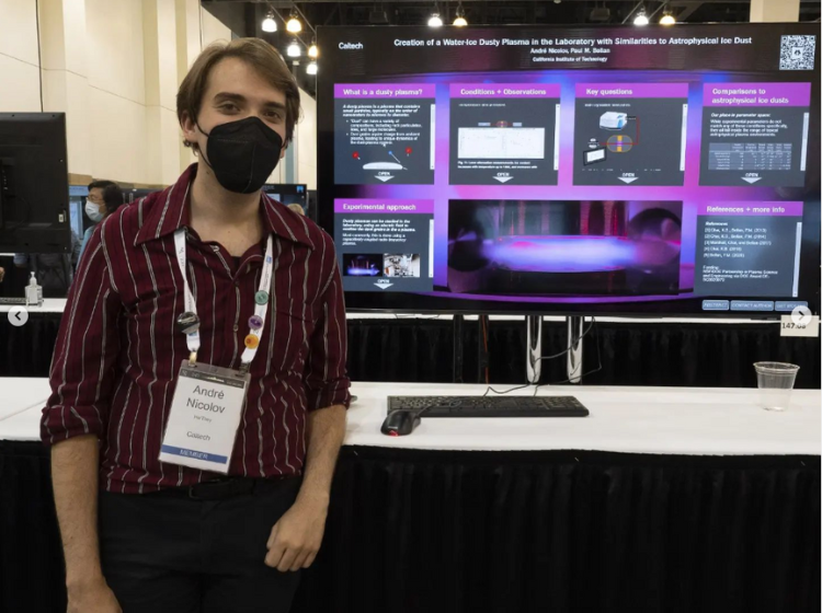 Presenter at AAS240 standing next to a large screen, facing the camera. On the screen is an iPoster with a bright purple, blue and black background. 