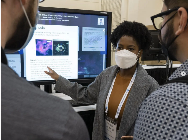 A Presenter at AAS240 standing next to a large screen and speaking to two other people. The presenter is pointing to an expanded content box on the screen while the 2 other people look on. 