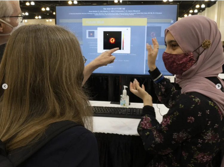 A Presenter standing next to a large screen and speaking to two other people. They are gesturing with their hands. On the screen is an iPoster with a content box expanded showing an image with a orange yellow orb against a black background. 