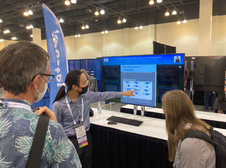 A Presenter at AAS240 standing next to a large screen and speaking to two other people. The presenter is pointing to an image of graphs on the screen while the 2 other people look on. 