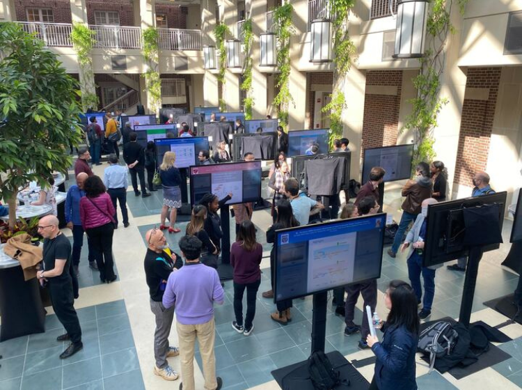 5 rows of 3 standing touch screens  displaying iPosters at the HHMI March Science Meeting. people are standing around, chatting and interacting with the screens. 