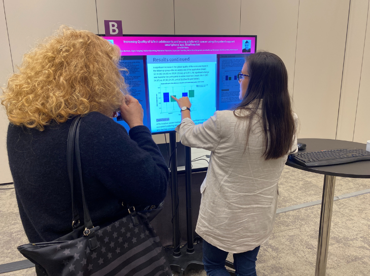 Two people standing in-front of an iPoster on a big screen at the ISOQOL annual conference.  They are facing towards the screen and one of them is pointing at an image on the screen.  