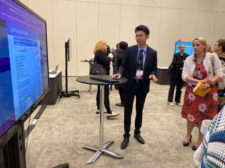 A person standing to the side of an iPoster on a big screen at the ISOQOL annual conference.  They are using a mouse on a 
small table to control the screen. Other people are crowded around in a semi circle watching the screen. 