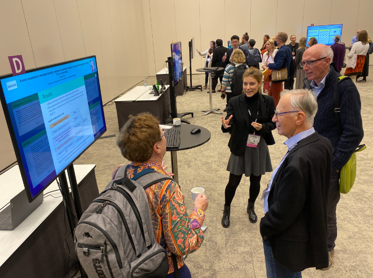 four people standing around an iPoster on a big screen at the ISOQOL annual conference. They appear to be discussing something. 