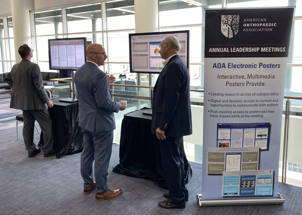 image from the  AOA event: 3 people standing inside at an event space. A stand up banner to the right advertises the American Orthopaedic Association Annual leadership meeting. There are 2 screens behind the people showcasing iPosters. 2 of the people are talking to one another in front of one screen and the 3rd is reading information on the other screen. 