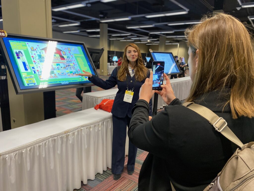 A Presenter posing next to an i-presentation on a screen whilst another person takes a picture of them. They are at the American Educational Research Association AERA 2023 Annual Meeting 