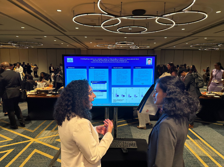 two people stand facing each other and talking in front of a screen with an iPoster displayed on it at the Skin of Color Society conference. 