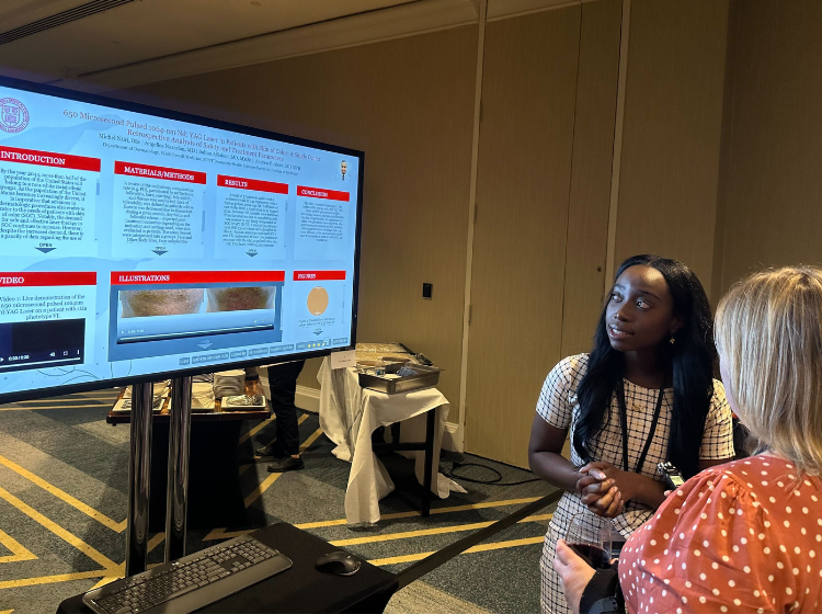 two people stand facing each other and talking in front of a screen with an iPoster displayed on it at the Skin of Color Society conference. 