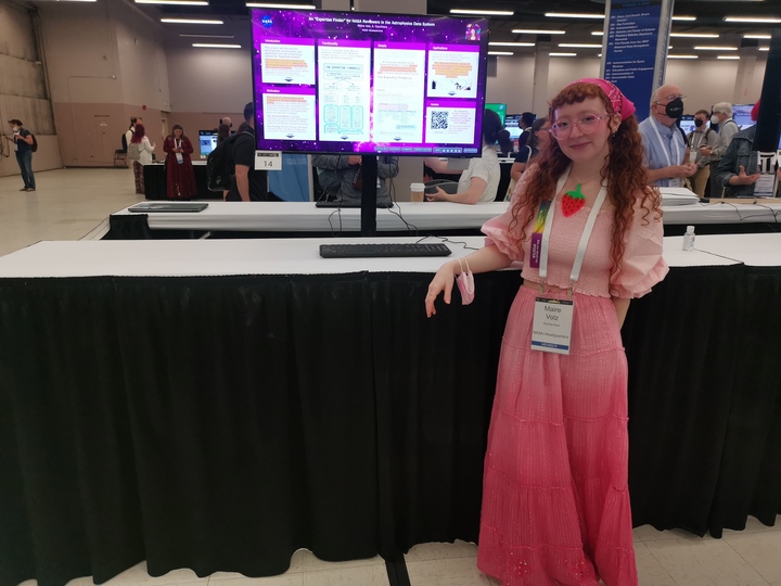 A presenter stands beside their iPoster on a standing screen at the AAS 242 summer meeting. 