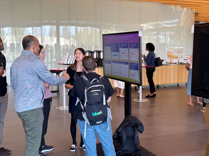 a group of people standing and interacting around an iPoster on a touch screen during a poster session at the Hana Gray Meeting. 