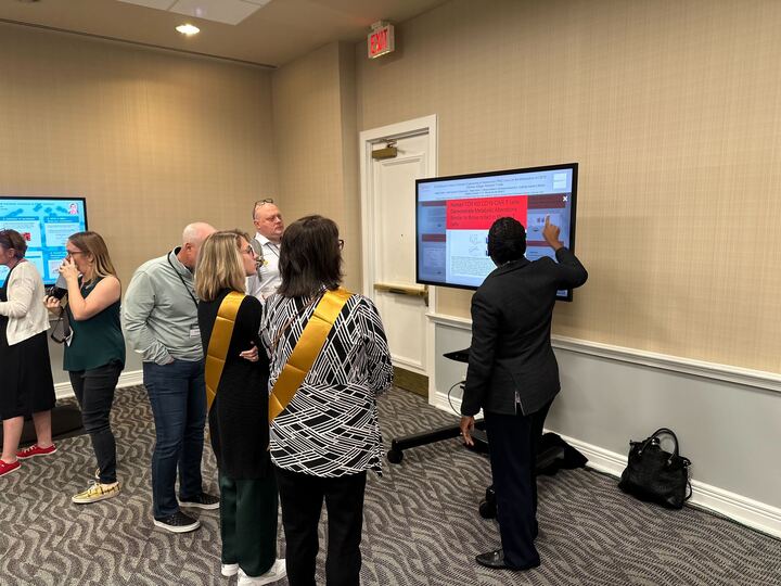A group of people standing around a touch screen with an iPoster displayed on it at the 2023 V Summit. One person is presenting the poster 