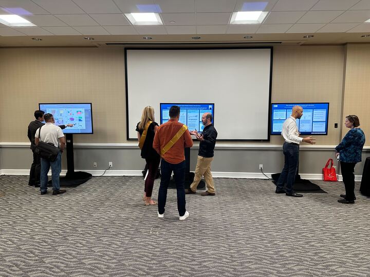 iPoster sessions taking place at the V Summit 2023. 3 touch screens display different iPosters. People are gathered in small groups around each one. 