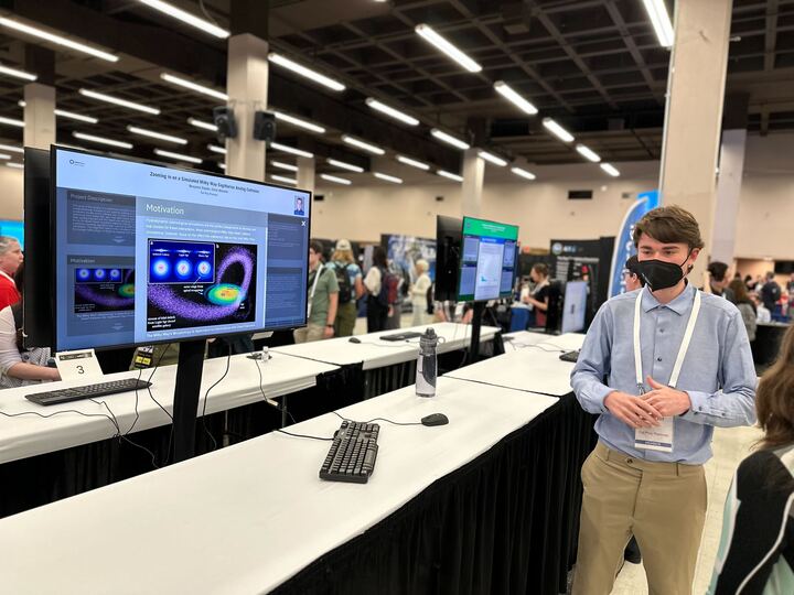 A presenter wearing a mask stands beside their iPoster on a standing screen at the AAS 242 summer meeting. 